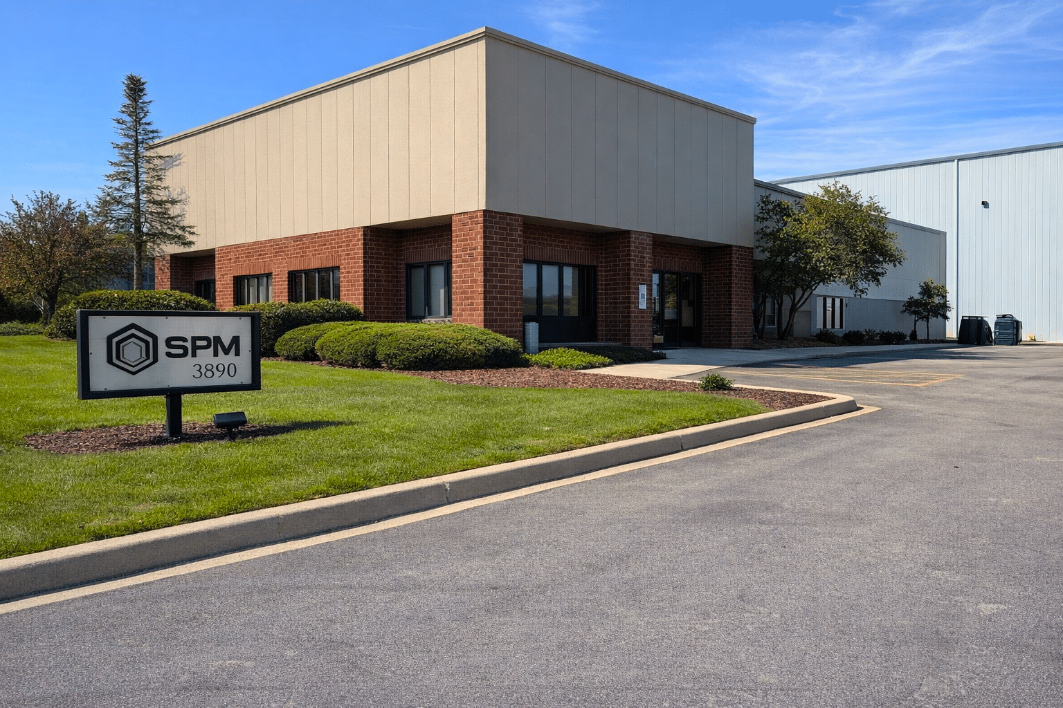 A commercial building with beige and red brick exterior sits beside a driveway. A sign in front reads SPM 3890. Green bushes, lawn, and a blue sky with light clouds are overhead.